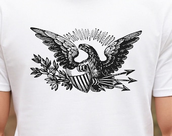 Puede incluir: Camiseta blanca con una ilustración en blanco y negro de un águila sosteniendo flechas y un escudo con rayas. El águila está rodeada de un sol radiante y una rama con hojas.