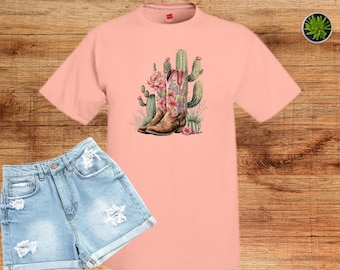 May include: A peach-colored short-sleeve t-shirt featuring a central graphic of brown cowboy boots adorned with pink flowers, surrounded by green cacti. The shirt is paired with light blue distressed denim shorts.