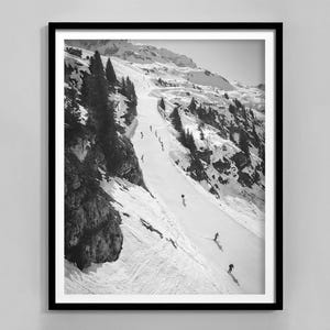 Op de afbeelding: Zwart-witfoto van een besneeuwde berghelling met skiërs die een piste afdalen. De skiërs zijn klein in de verte, en de berg is bedekt met sneeuw en bomen.
