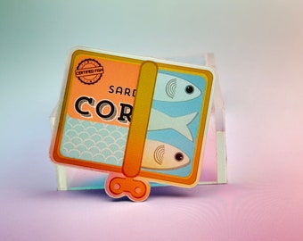 May include: A yellow and blue sticker with a can of sardines. The can has a label that says "CERTIFIED FISH" and "SARDINES CORTE", with a pattern of blue and white scales. The can has two sardines inside, one on top of the other. The sticker has a yellow pull tab.
