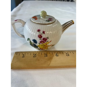 Puede incluir: Una tetera de porcelana blanca con cuerpo acanalado, decorada con motivos florales coloridos y una tapa con borde dorado y pomo verde pálido. Una regla de madera colocada delante muestra una anchura total de unos 14 cm.