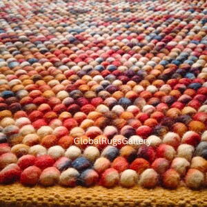 Op de afbeelding: Een kleurrijk tapijt met een textuur, met een patroon van kleine, ronde, vervilte wollen ballen. Het kleurenpalet van het tapijt omvat tinten rood, oranje, geel, blauw en wit. Het tapijt is een decoratieve vloerbedekking.
