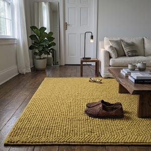 Puede incluir: Una alfombra amarilla mostaza con textura es el punto focal de una sala de estar. Zapatos de cuero marrón y gafas están sobre la alfombra. También se ven una mesa de centro de madera, un sofá y una lámpara negra.