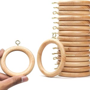 May include: A stack of light-colored wooden curtain rings with brass-colored hooks. One ring is held by a hand, and another is positioned in front of the stack. The rings have a smooth, natural finish and are designed for hanging curtains.