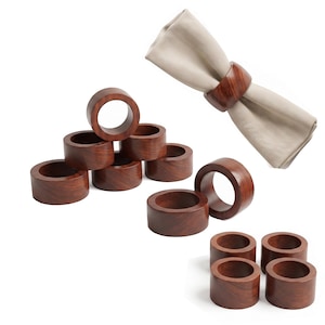 May include: A set of circular wooden napkin rings with a polished reddish-brown wood grain finish. One ring is shown holding a folded beige fabric napkin. The rings are arranged in a group, displaying their smooth, cylindrical, and hollow design.