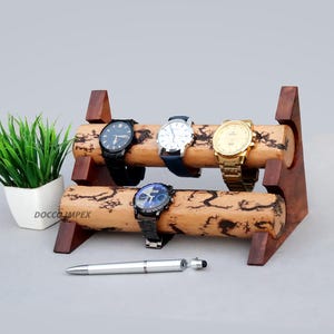 Puede incluir: Un expositor de relojes de madera con cuatro relojes de pulsera con correas y esferas de diferentes colores. El expositor es de madera oscura y tiene un aspecto natural y rústico. Un bolígrafo y una pequeña planta en maceta también están en la imagen.