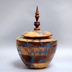 Op de afbeelding: Een handgemaakte houten urn met een deksel en een decoratieve finial. De urn heeft een natuurlijk houtnerfpatroon met blauwe harsaccenten. De urn is ongeveer 25 cm hoog.