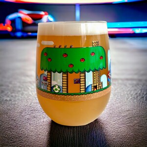 May include: A clear glass beer stein with a yellow, green, and brown pixelated design of a video game level. The design features a green tree with red mushrooms, a brown house, and a yellow sign that reads "Yoshi".