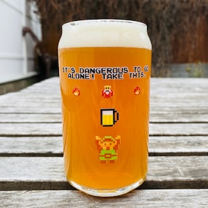 May include: A glass of beer with a white head. The glass has a graphic of a video game character and the text "It's dangerous to go alone! Take this."