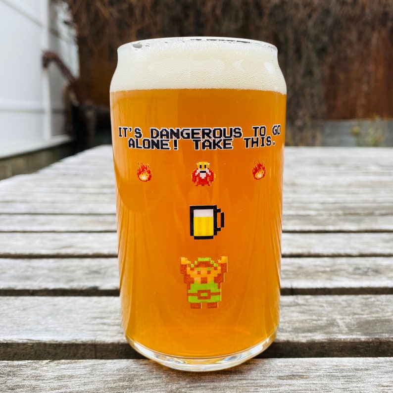 May include: A glass of beer with a white head. The glass has a graphic of a video game character and the text "It's dangerous to go alone! Take this."