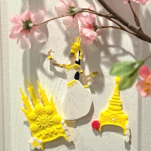 May include: A decorative set featuring a white and yellow figurine of a traditional dancer, along with yellow headpieces. Pink cherry blossoms and a branch are in the background. The dancer is wearing a white dress and yellow accessories.