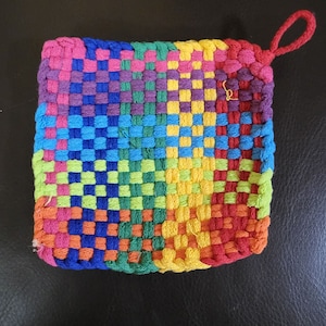 May include: A colourful woven pot holder with a loop handle. The pot holder is made of different coloured yarn, including red, orange, yellow, green, blue, and purple.