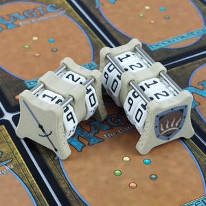 May include: Two beige and white dice counters for a tabletop game. Each counter has a rotating cylinder with numbers 0-9 and 1-2. One counter has a sword design, the other a shield. The counters are on a game mat.
