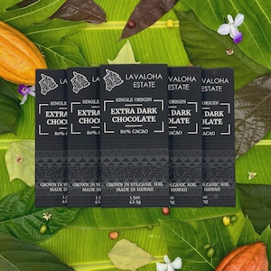 May include: Several dark chocolate bars with the text "LAVALOHA ESTATE" and "EXTRA DARK CHOCOLATE 80% CACAO" are arranged on a bed of green leaves. The bars are 1.5 oz (42.5g) each. The image includes cacao pods and flowers.