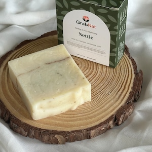 May include: A bar of natural handmade soap with a green and white label that reads "GrabNat Healing & Pore Tightening Nettle 100% All-Natural Handmade Soap" on a wooden slice.
