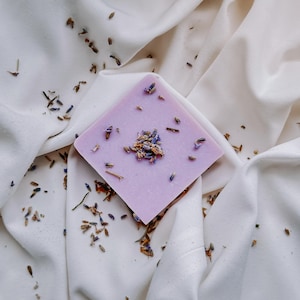 May include: A square bar of lavender soap with dried lavender flowers on top. The soap is a light purple colour and is sitting on a white fabric background.