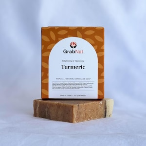 Może przedstawiać: Kostka mydła z kurkumą z pudełkiem na wierzchu. Pudełko jest pomarańczowe z białą etykietą z napisem "GrabNat Turmeric" i "100% ALL-NATURAL HANDMADE SOAP". Mydło ma jasnobrązowy kolor.