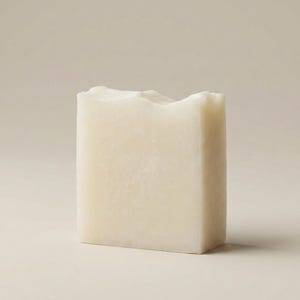 Peut inclure: Un pain de savon rectangulaire blanc cassé avec un bord supérieur texturé et irrégulier. Le savon a un design simple et minimaliste, et est posé sur un fond neutre. Le savon semble fait à la main.