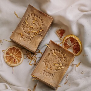 May include: Two brown bars of soap with a floral design and the word "TOP" embossed on them. The soap is on a white fabric background with dried orange slices and flower petals.