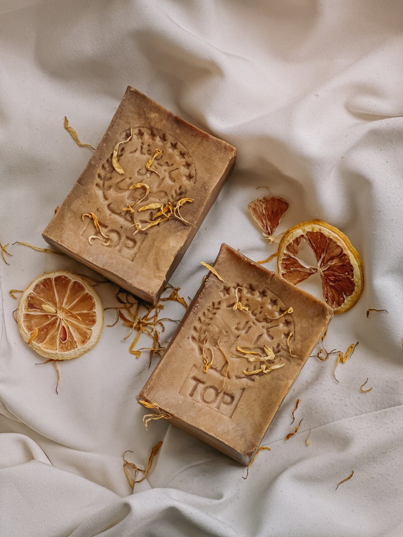 May include: Two brown bars of soap with a floral design and the word "TOP" embossed on them. The soap is on a white fabric background with dried orange slices and flower petals.