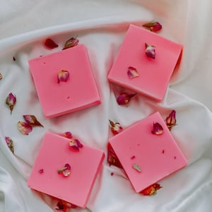 May include: Four pink square-shaped soaps with dried rose petals on top, arranged on a white fabric background.