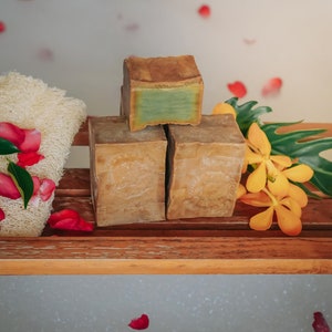 May include: Three bars of natural soap, two brown and one green, on a wooden shelf with a white bath tub in the background.  Rose petals are floating in the bath water.