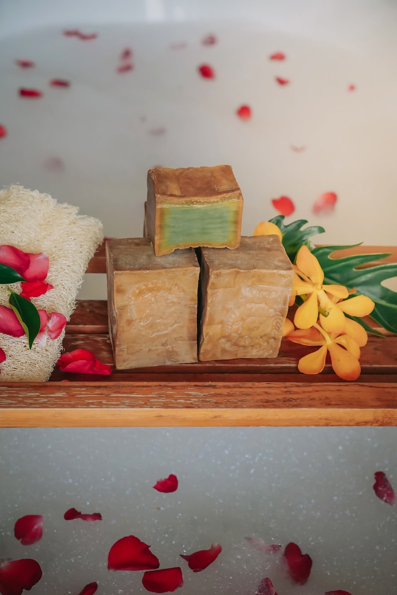 May include: Three bars of natural soap, two brown and one green, on a wooden shelf with a white bath tub in the background.  Rose petals are floating in the bath water.