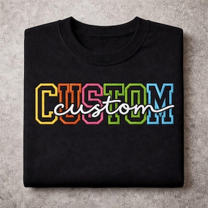 Puede incluir: Sudadera negra con la palabra "CUSTOM" en un diseño de letras de bloque colorido. La palabra "custom" está en una fuente cursiva blanca y se coloca sobre las letras de bloque. La sudadera está doblada sobre una superficie gris.