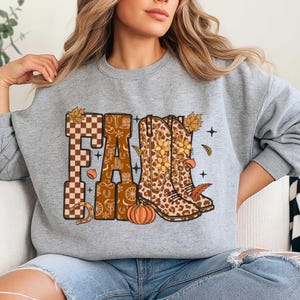 May include: A gray sweatshirt with a fall-themed graphic. The word "FALL" is displayed in a mix of patterns, including a checkerboard and a brown floral design. The graphic also features cowboy boots, a pumpkin, and autumn leaves.