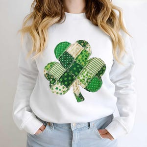 May include: White sweatshirt featuring a large, patchwork shamrock design. The shamrock is composed of various green fabrics with patterns like polka dots, stripes, and floral prints. The sweatshirt has long sleeves and a crew neck.