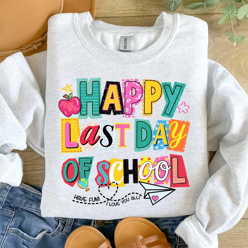 K&ouml;nnte beinhalten: Ein hellgraues Sweatshirt mit der Aufschrift "HAPPY LAST DAY OF SCHOOL" in bunten Blockbuchstaben. Das Design umfasst einen Apfel, einen Globus, ein Papierflugzeug und die S&auml;tze "HAVE FUN!" und "I LOVE YOU ALL!"