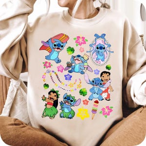May include: Beige sweatshirt featuring a colorful graphic of the character Stitch from Lilo & Stitch. The design includes Stitch with a surfboard, ice cream, and other tropical elements. The phrase "OHANA MEANS FAMILY" is also included.