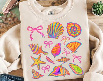 Op de afbeelding: Een beige sweatshirt met een kleurrijk schelp- en zeestermotief. Het ontwerp bevat roze strikjes, schelpen in tinten geel, oranje en roze, en een zeester in geel, orange en roze.