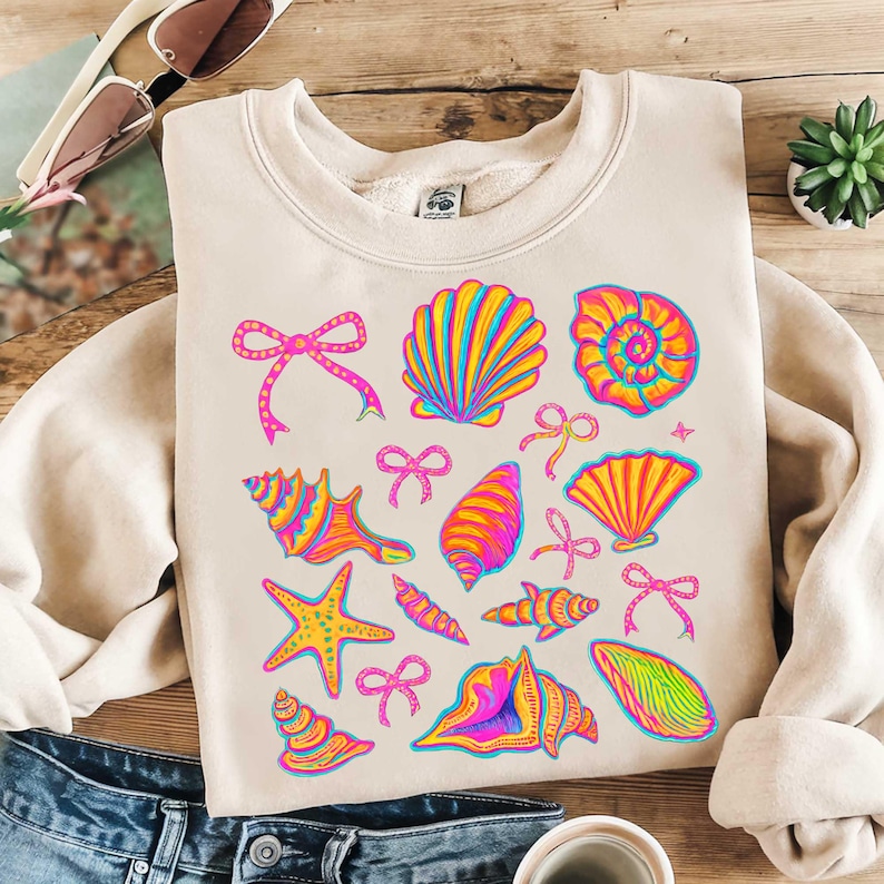May include: A beige sweatshirt with a colorful seashell and starfish pattern. The design features pink bows, seashells in shades of yellow, orange, and pink, and a starfish in yellow, orange, and pink.