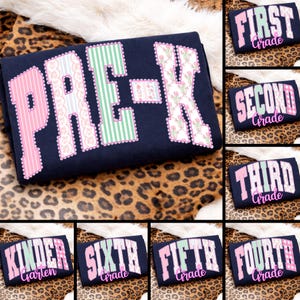 Puede incluir: Camiseta azul marino de Pre-K con letras estampadas en rosa y verde. Camisetas adicionales muestran "First Grade", "Second Grade", "Third Grade", "Kindergarten", "Sixth Grade", "Fifth Grade" y "Fourth Grade".