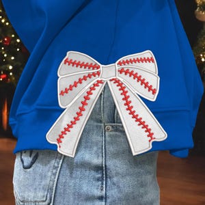 May include: A blue sweatshirt with a white and red baseball-themed bow on the back. The bow is made of white fabric with red stitching that resembles baseball stitching.