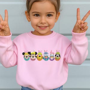 Op de afbeelding: Een lichtroze sweatshirt met een paasontwerp. Het ontwerp bevat zes cartoon-personage-eieren, elk met een ander gezicht en konijnenoren. De sweatshirt heeft een ronde hals en lange mouwen.