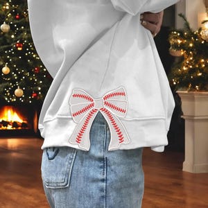 May include: White sweatshirt with a red and white baseball-themed bow on the back.