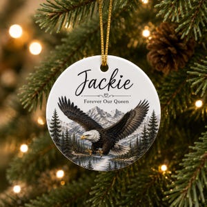 Op de afbeelding: Een rond wit keramieken ornament dat aan een gouden koord hangt. Het ornament toont een Amerikaanse zeearend in vlucht boven een meer, dennenbomen en besneeuwde bergen. De tekst luidt Jackie, gevolgd door een hartje en Forever Our Queen.