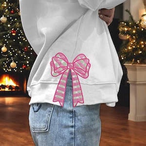 May include: A white sweatshirt with a pink and green plaid bow design. The bow is embroidered on the lower back of the sweatshirt. The sweatshirt is paired with blue jeans.