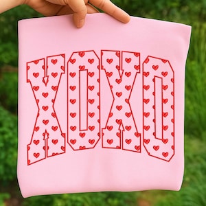 May include: A light pink sweatshirt with the letters "XOXO" in a bold, red, collegiate-style font. Each letter is filled with a pattern of small red hearts, creating a playful and romantic design.