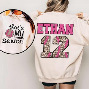 Könnte beinhalten: Cremefarbenes Sweatshirt mit dem Namen "ETHAN" in rosa Blockbuchstaben und der Zahl "12" in einem Leopardenmuster mit rosa Baseballnähten. Auf der Vorderseite befindet sich ein kreisförmiges Design mit dem Text "that's MY Senior!" und einem Baseball.