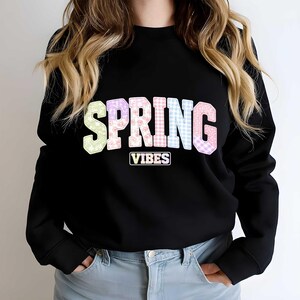 Op de afbeelding: Zwarte sweatshirt met de tekst "SPRING VIBES" in pastelkleuren. Het woord "SPRING" heeft een patchwork-ontwerp met bloemen- en ruitpatronen. "VIBES" staat in een witte rechthoek.