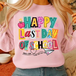 K&ouml;nnte beinhalten: Ein hellrosa Sweatshirt mit der Aufschrift "HAPPY LAST DAY OF SCHOOL" in bunten Blockbuchstaben. Das Design umfasst einen Apfel, einen Globus, ein Papierflugzeug und die S&auml;tze "HAVE FUN!" und "I LOVE YOU ALL!"