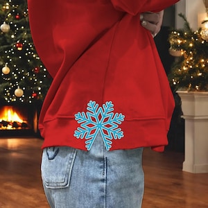 May include: A red sweatshirt with a light blue snowflake design on the lower back. The snowflake has a detailed outline and a lighter blue center. The sweatshirt is paired with blue jeans.