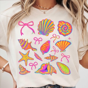 May include: A white t-shirt with a colorful seashell and starfish design. The design features pink, yellow, blue, and green seashells, starfish, and bows.