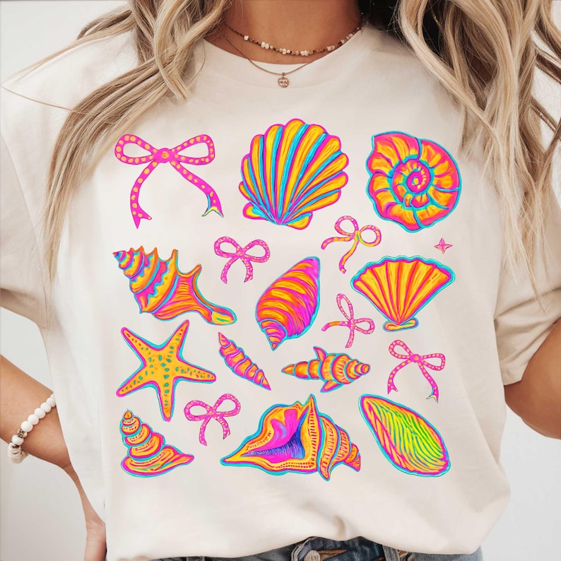 May include: A white t-shirt with a colorful seashell and starfish design. The design features pink, yellow, blue, and green seashells, starfish, and bows.