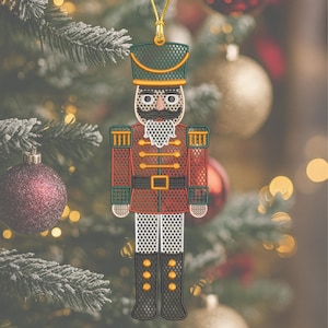 Könnte beinhalten: Gestickter Weihnachtsschmuck in Form eines Nussknacker-Soldaten. Der Schmuck hat eine rote Jacke, einen grünen Hut und schwarze Stiefel. Er hat eine goldene Aufhängeschlaufe und ist mit goldenen und weißen Details verziert. Der Hintergrund ist ein verschwommener Weihnachtsbaum.