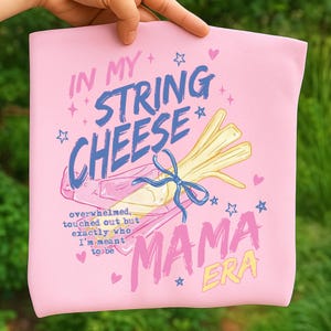Könnte beinhalten: Rosa T-Shirt mit dem Text "IN MY STRING CHEESE MAMA ERA". Das Design beinhaltet eine Illustration von Fädenkäse, der mit einem blauen Band, Sternen und Herzen gebunden ist. Der Text "overwhelmed, touched out but exactly who I'm meant to be" ist ebenfalls enthalten.