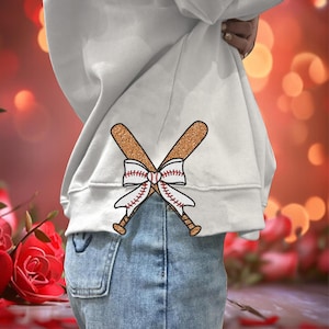 May include: White sweatshirt with a baseball bat bow design. The bats are crossed and tied together with a white ribbon with red stitching. The sweatshirt is paired with blue jeans.
